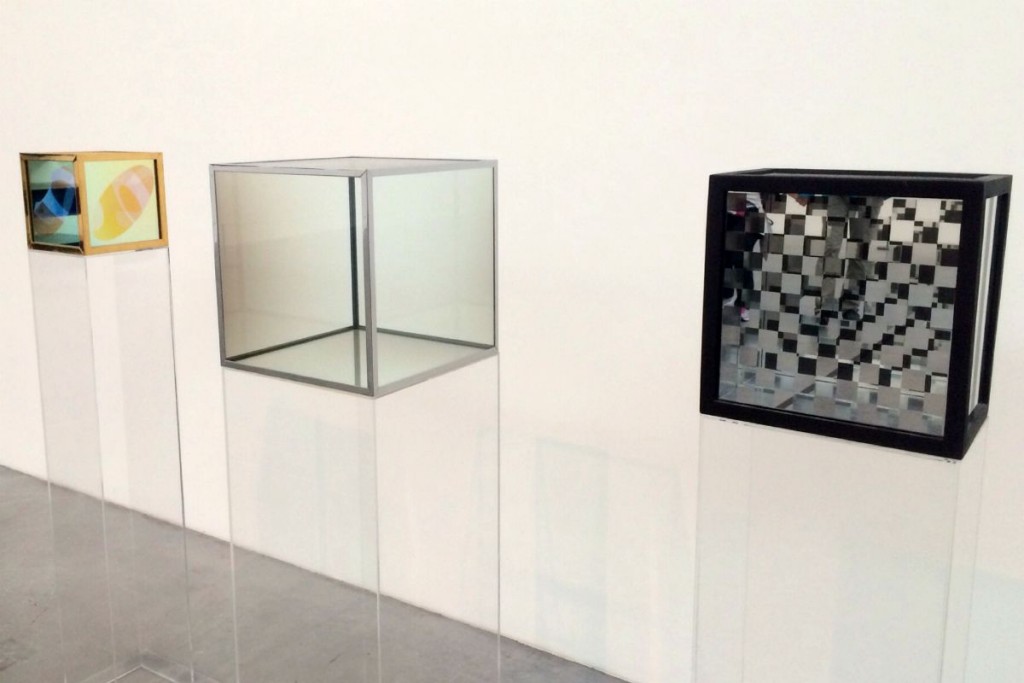 Artwork: three glass cubes sitting on glass columns. One has a gold frames and yellow abstract shapes on the sides, one has a silver frame and clear sides, and one has a back frame and checkrbaord sides.